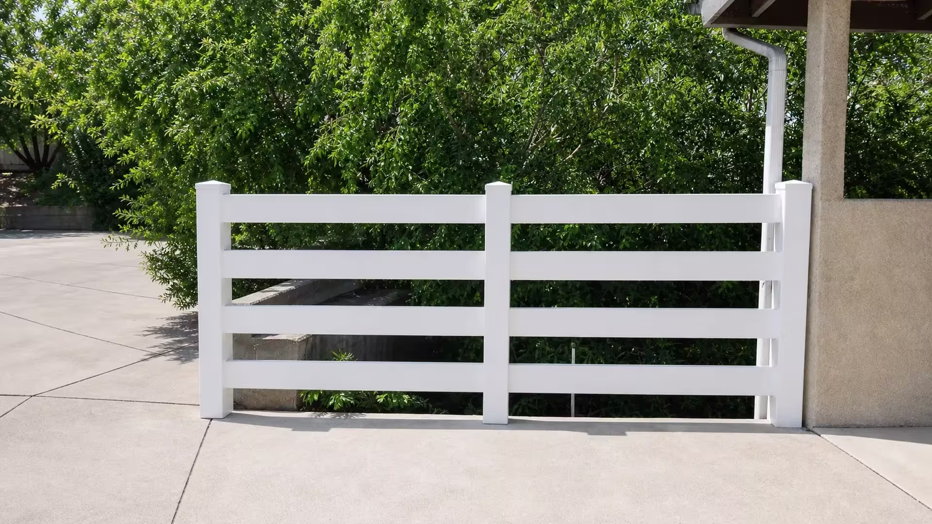 A stucco building's roof edge and downspout stand where the White vinyl rails of this ranch fence come to an end