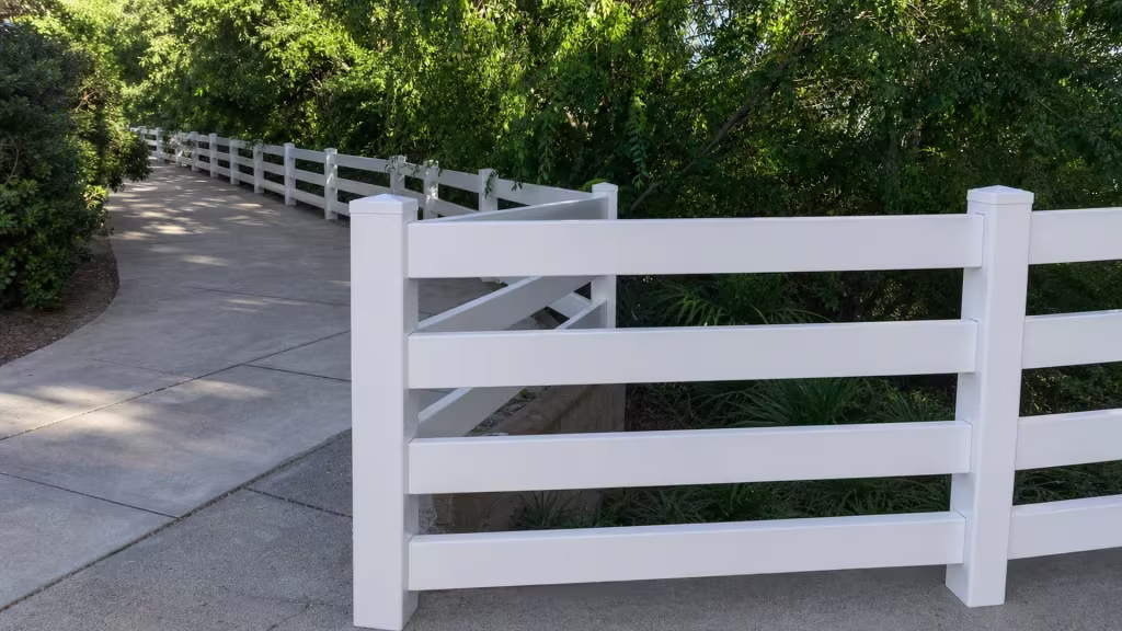 A low wooden retaining edge holds a descending planted slope beneath these White vinyl ranch fence rails