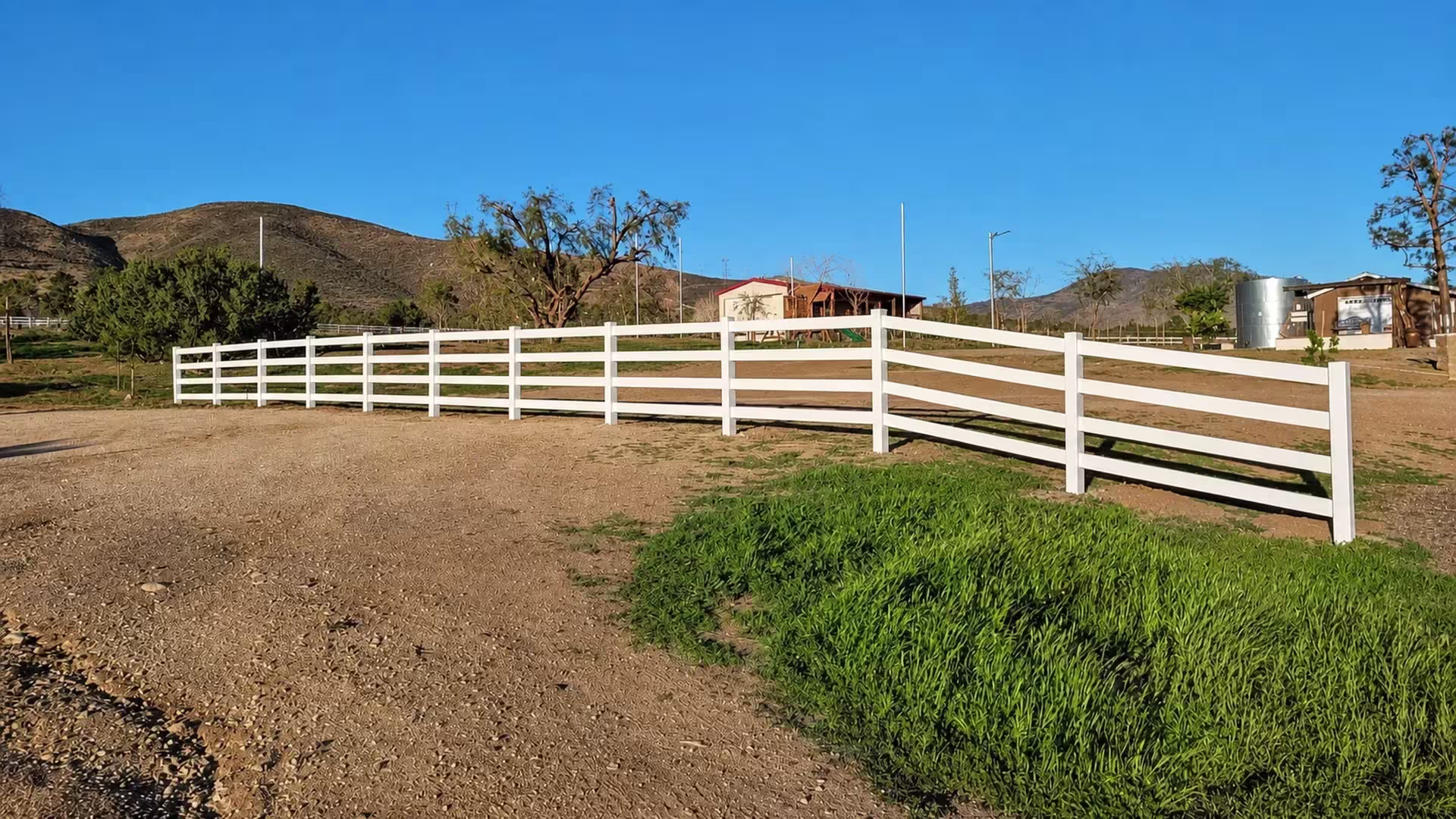 Dirt and gravel driveway ground carries a sweeping, turning run of White vinyl rails toward distant hills