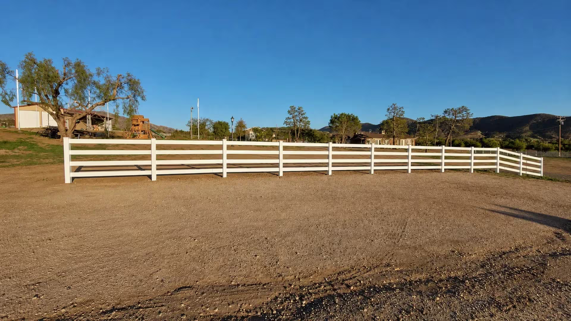 Sparse lawn and mature trees border the corner where this White vinyl fence's rails turn into a return run