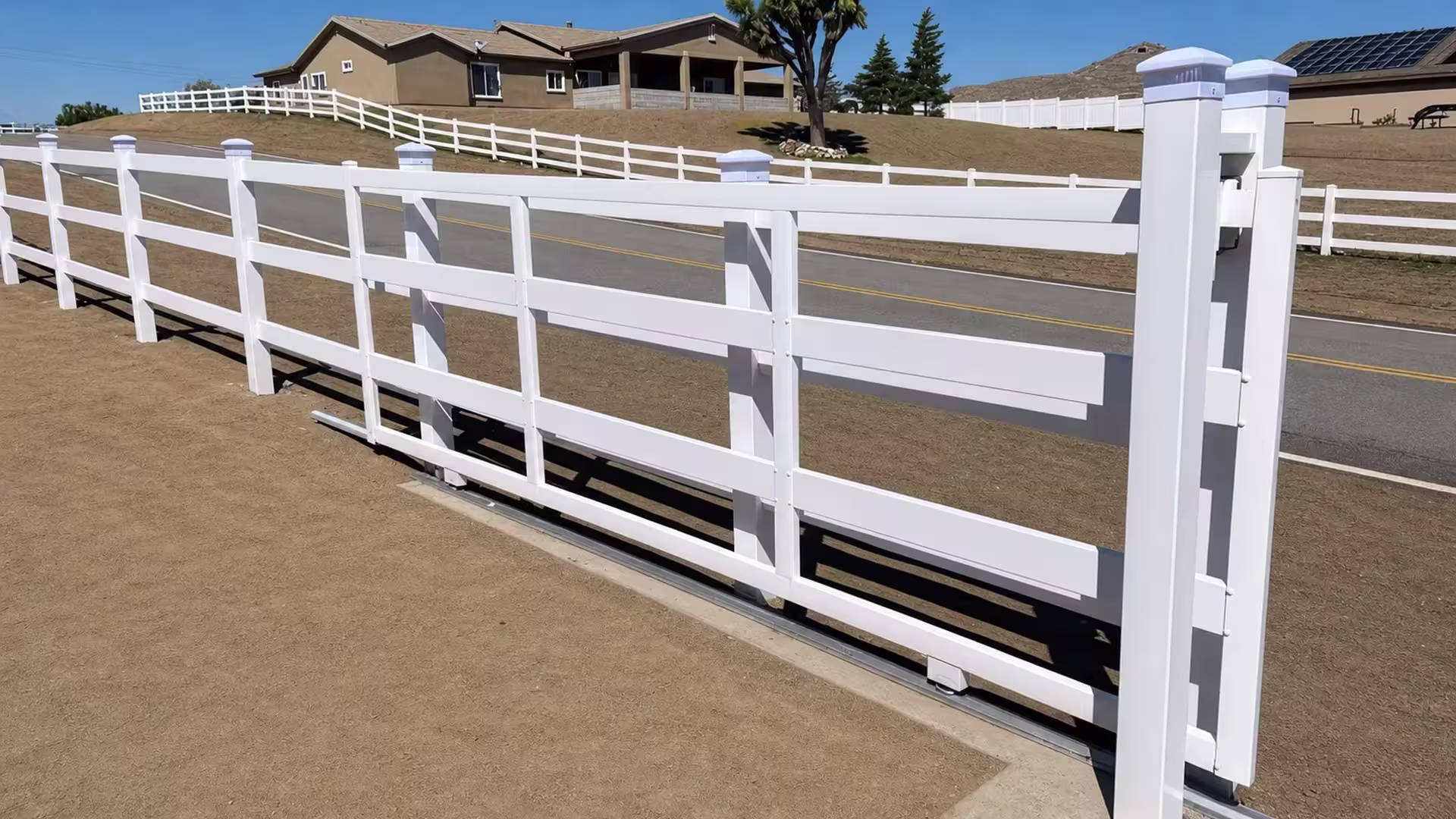 A metal sliding track sits beneath this White vinyl four-rail fence beside a paved road