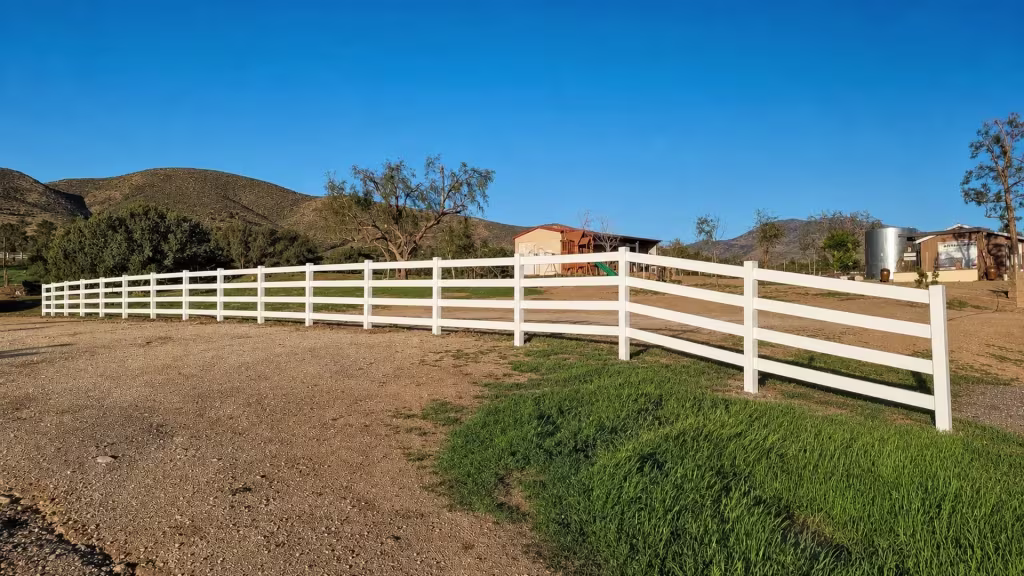 Utility poles and open hills rise behind this White vinyl ranch fence's four rails and gravel drive
