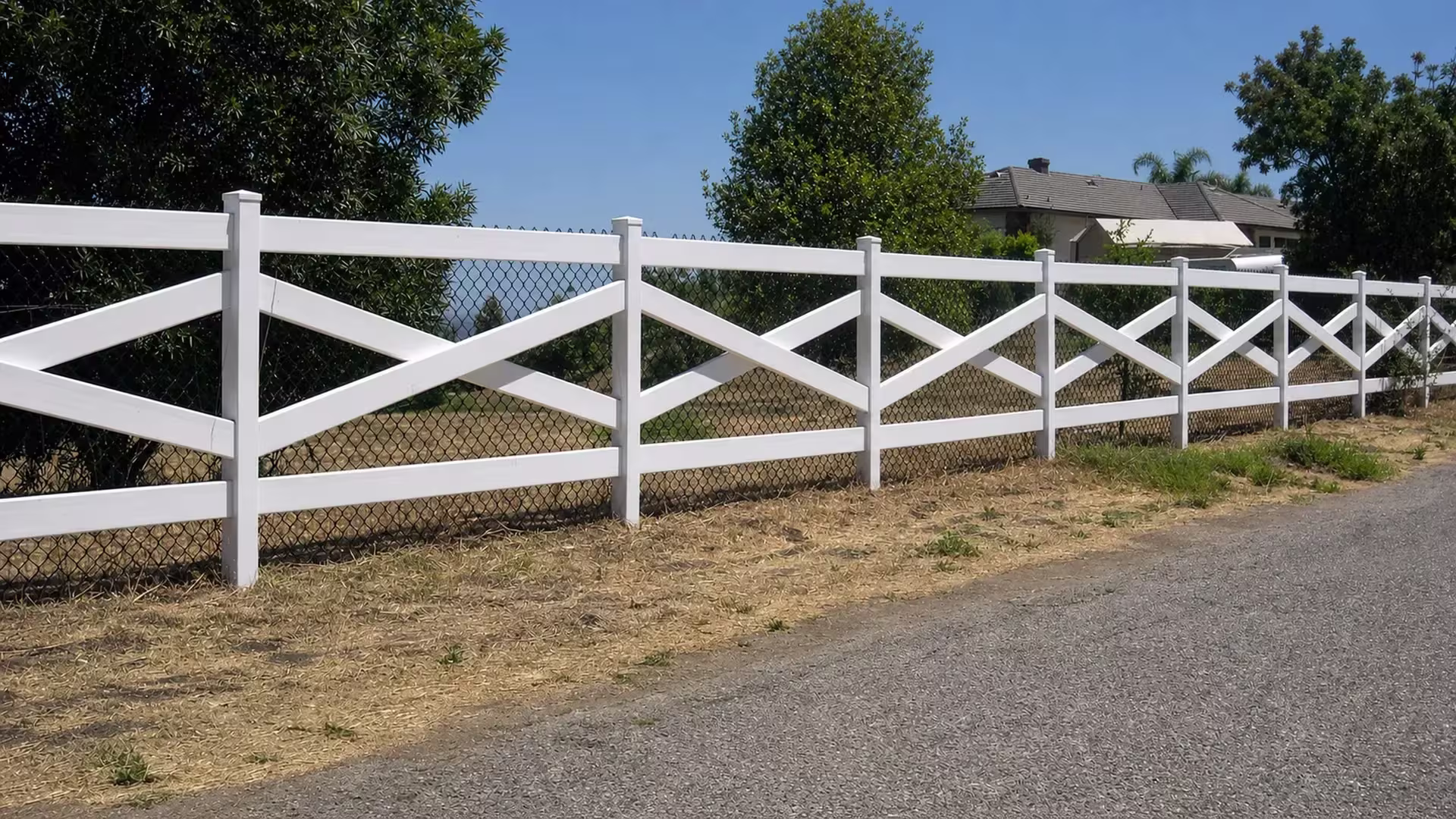 Dry soil and mature trees border a roadside verge tracked by this cross rail White vinyl fence