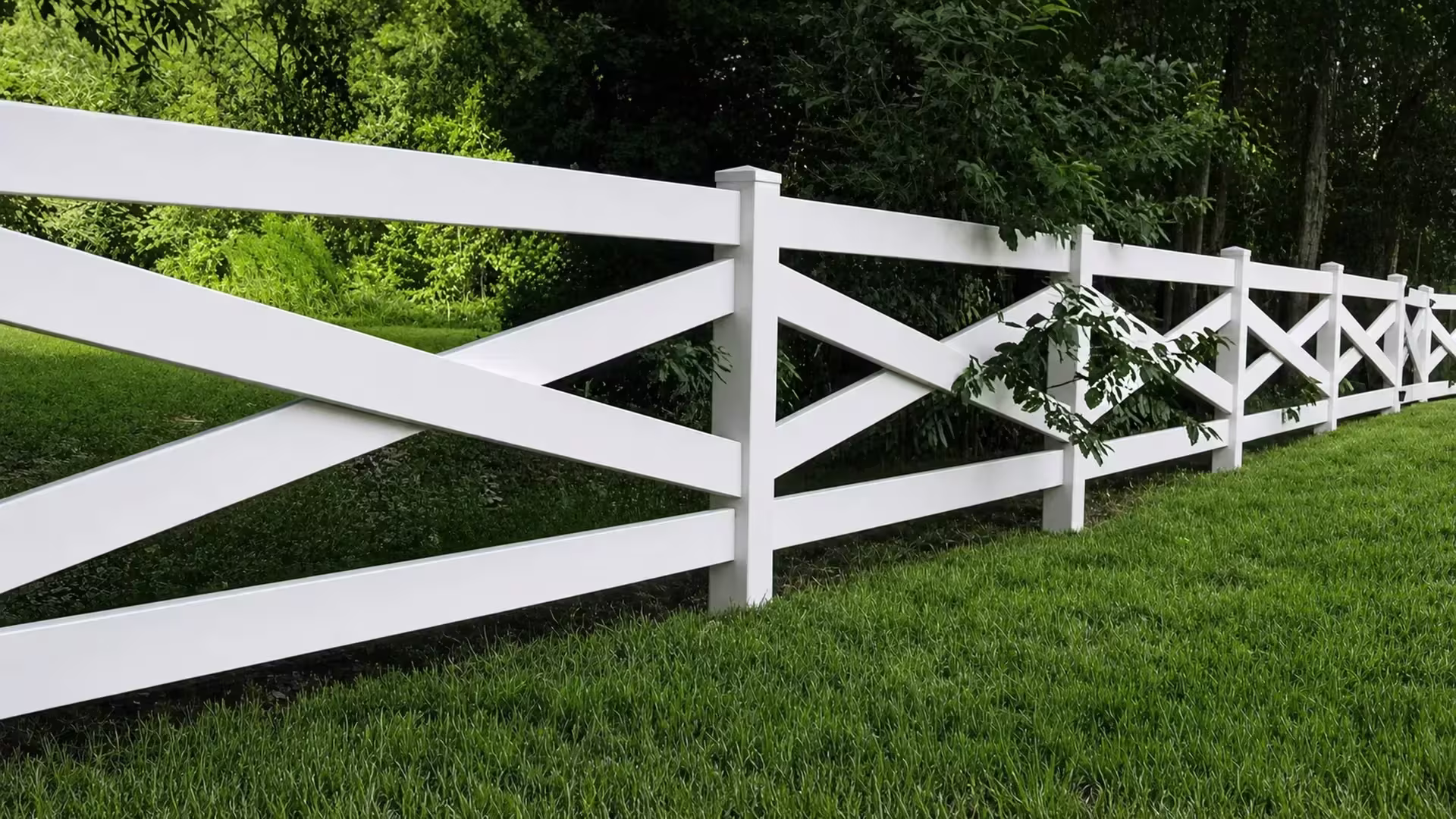 A leafy branch overlaps the bays of this cross rail White vinyl fence above a grassy hillside
