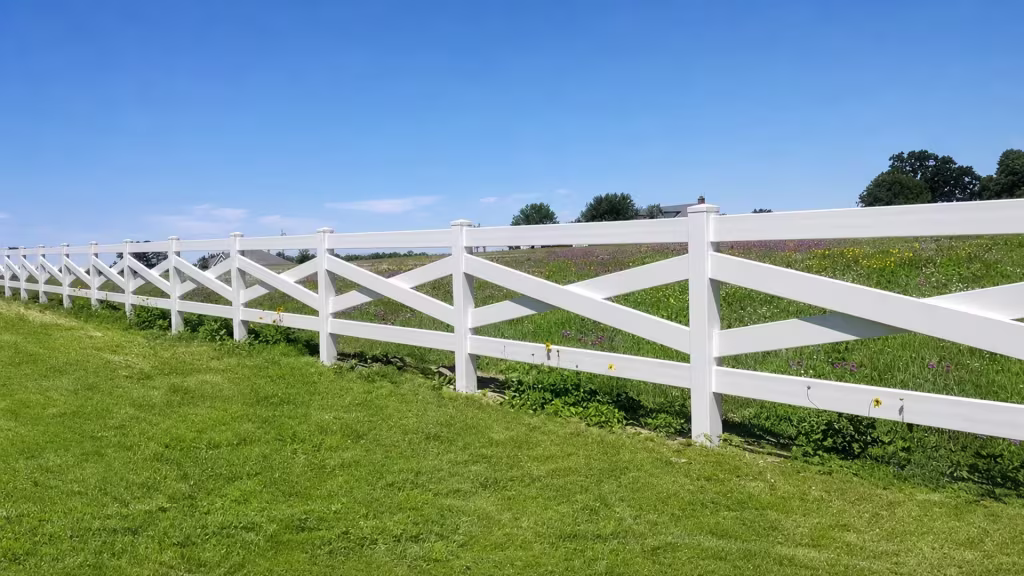 Eleven square posts recede in perspective along this cross rail White vinyl fence at the edge of a field