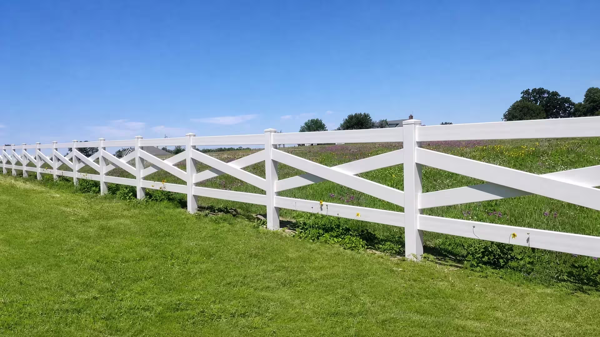 Eleven square posts recede in perspective along this cross rail White vinyl fence at the edge of a field