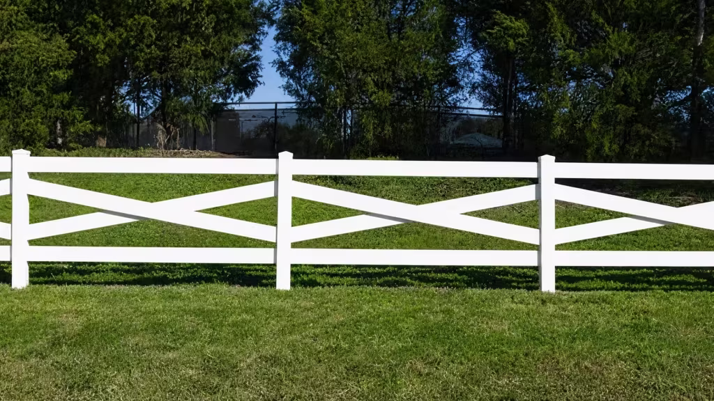 Three square posts and their pyramidal caps front a dark chain-link boundary behind this White vinyl cross-rail fence