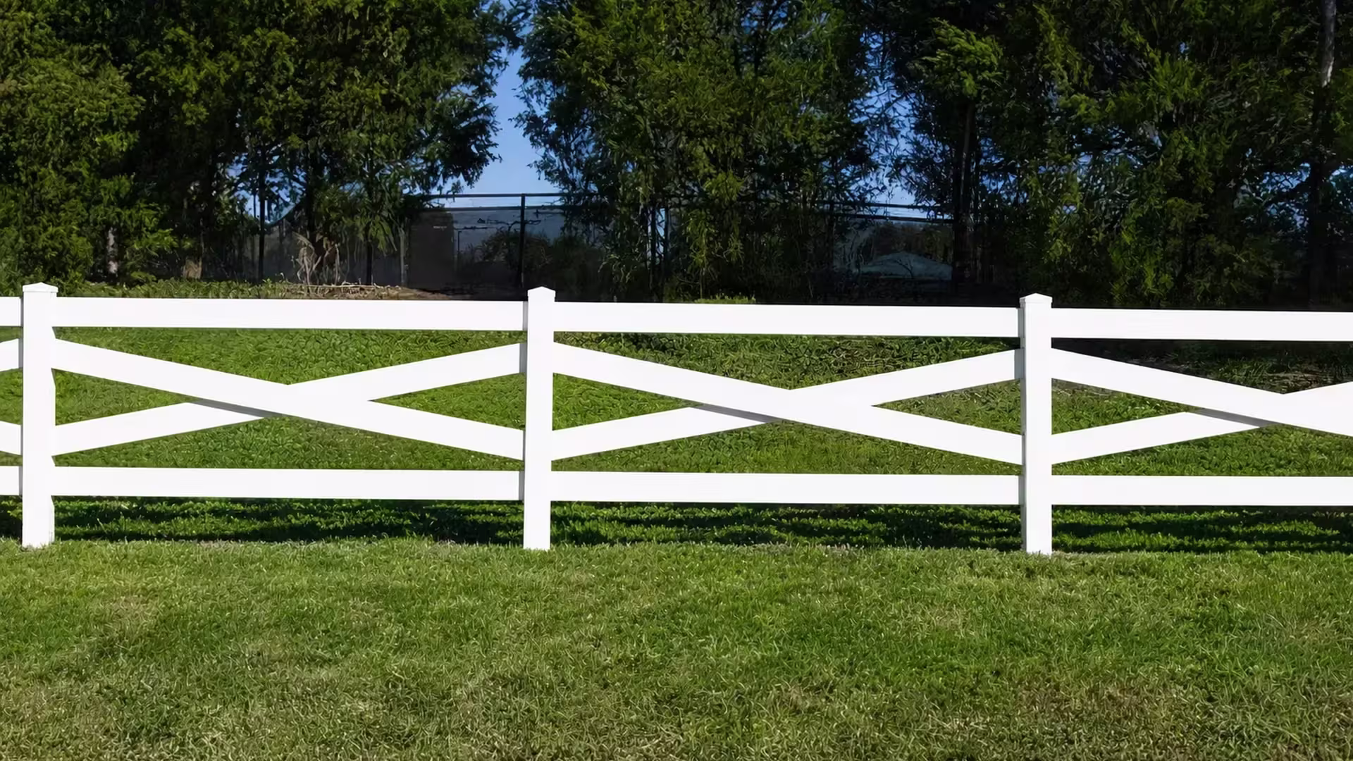 Three square posts and their pyramidal caps front a dark chain-link boundary behind this White vinyl cross-rail fence