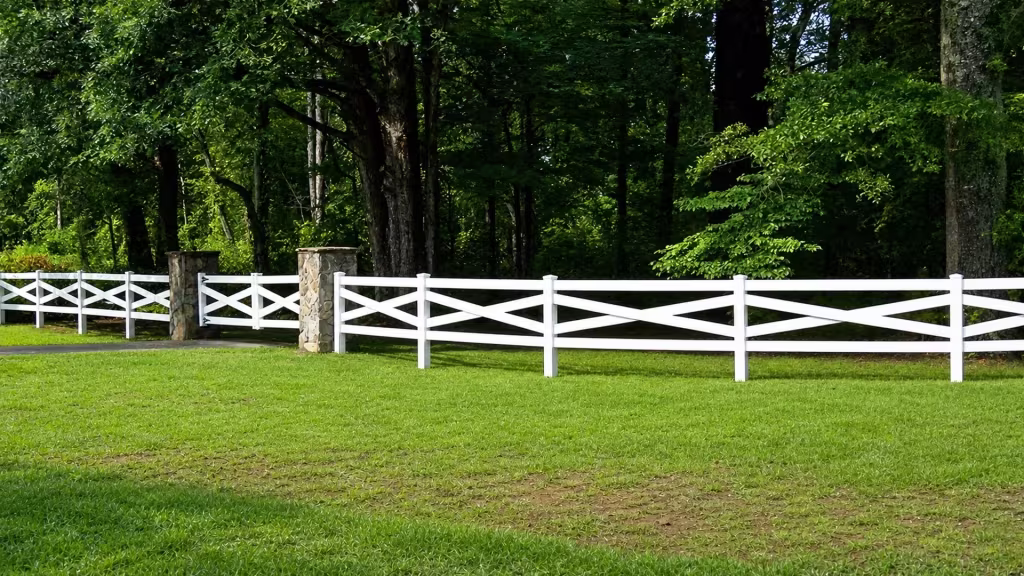 A masonry column and a paved strip sit beside this cross rail White vinyl fence in a wooded lawn
