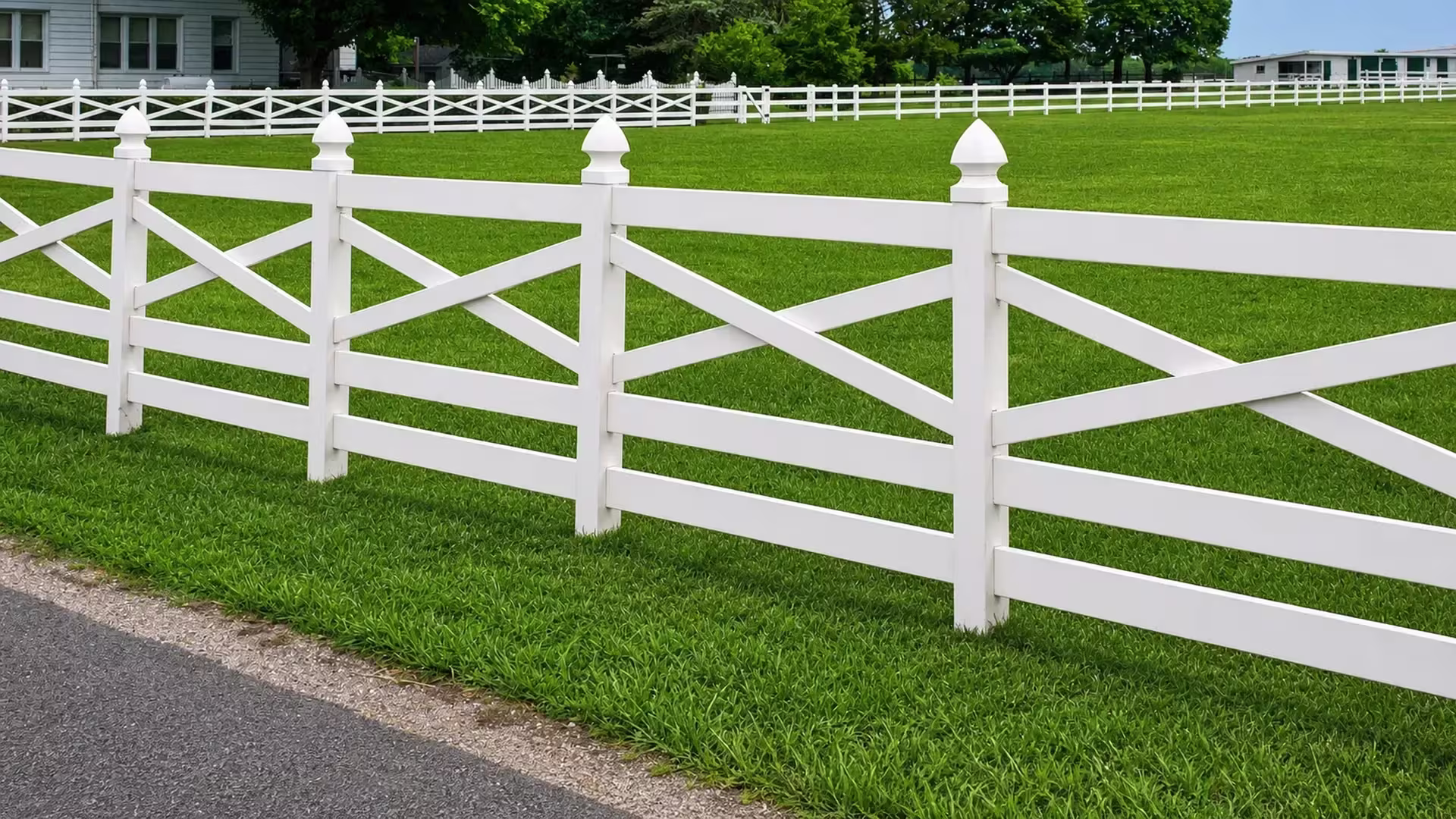 An asphalt edge runs past a house and a distant building beside this cross rail White vinyl fence