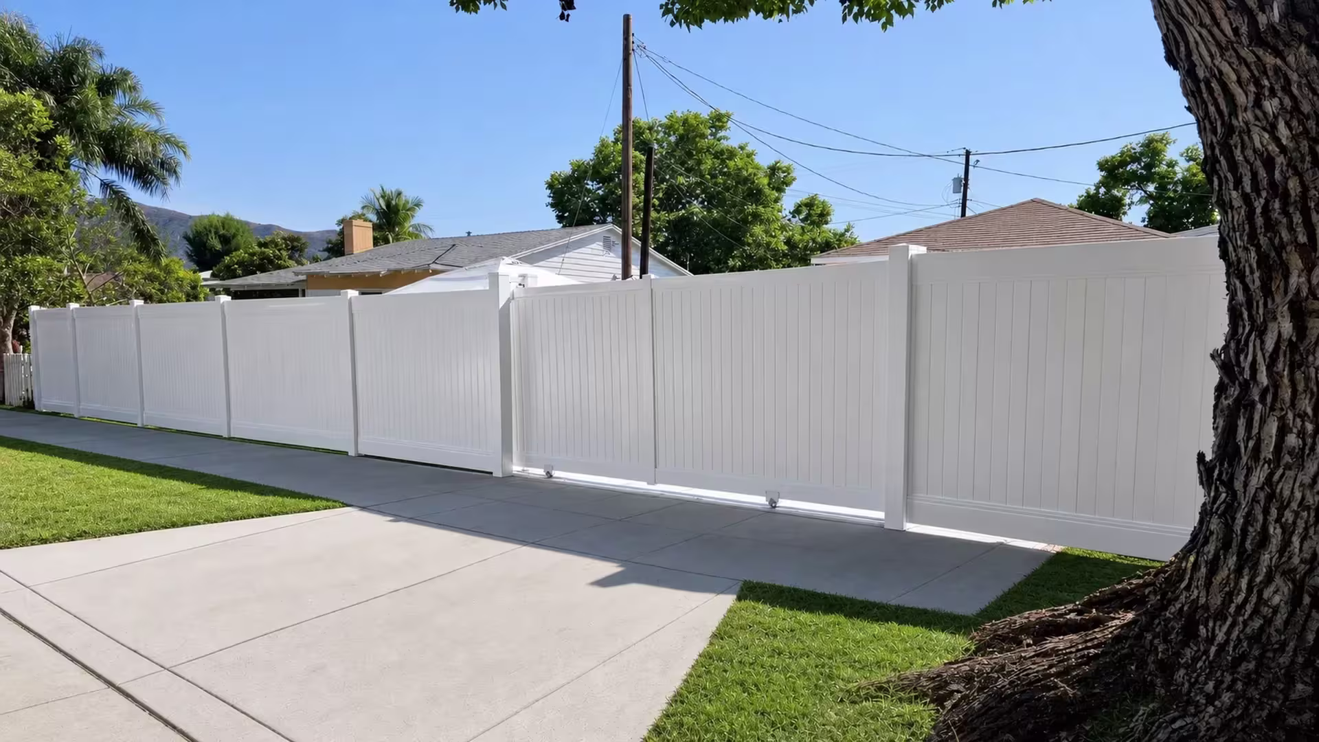 White vinyl privacy rolling gate integrated into a stepped fence line, framed by a large rooted tree and a distant mountain
