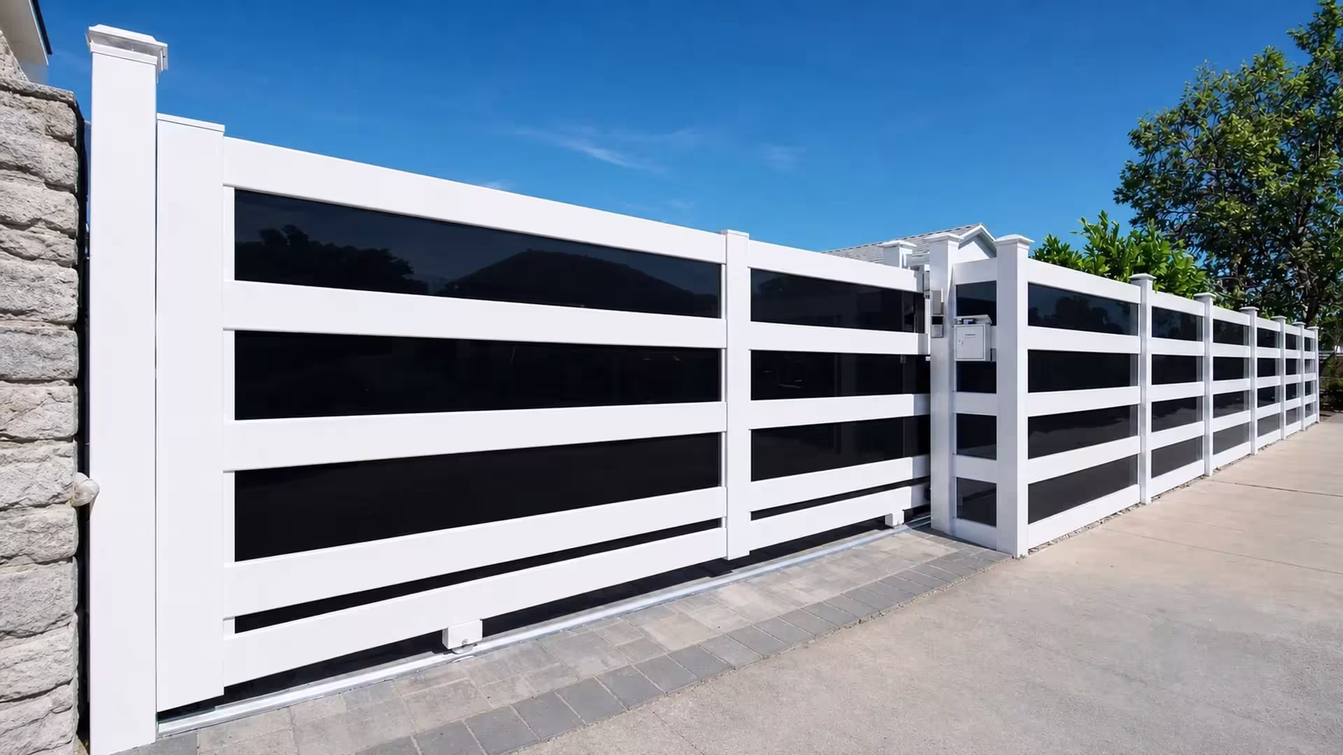 White vinyl rolling gate with smoke-black acrylic infill starting beside a stone wall and a gray-and-tan paver strip inset