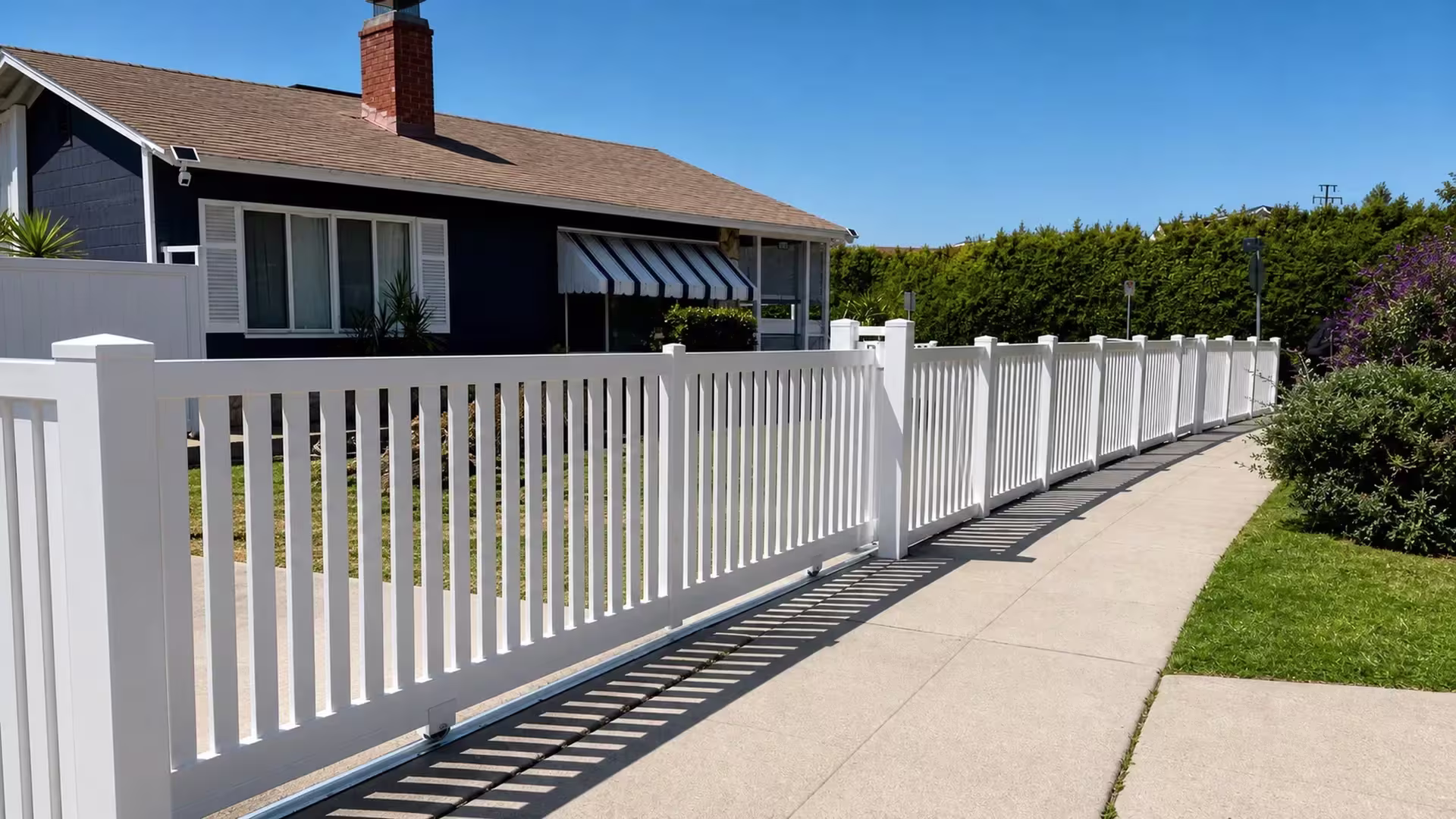 White vinyl rolling picket gate sliding on a ground track in front of a house with a brick chimney and striped awning