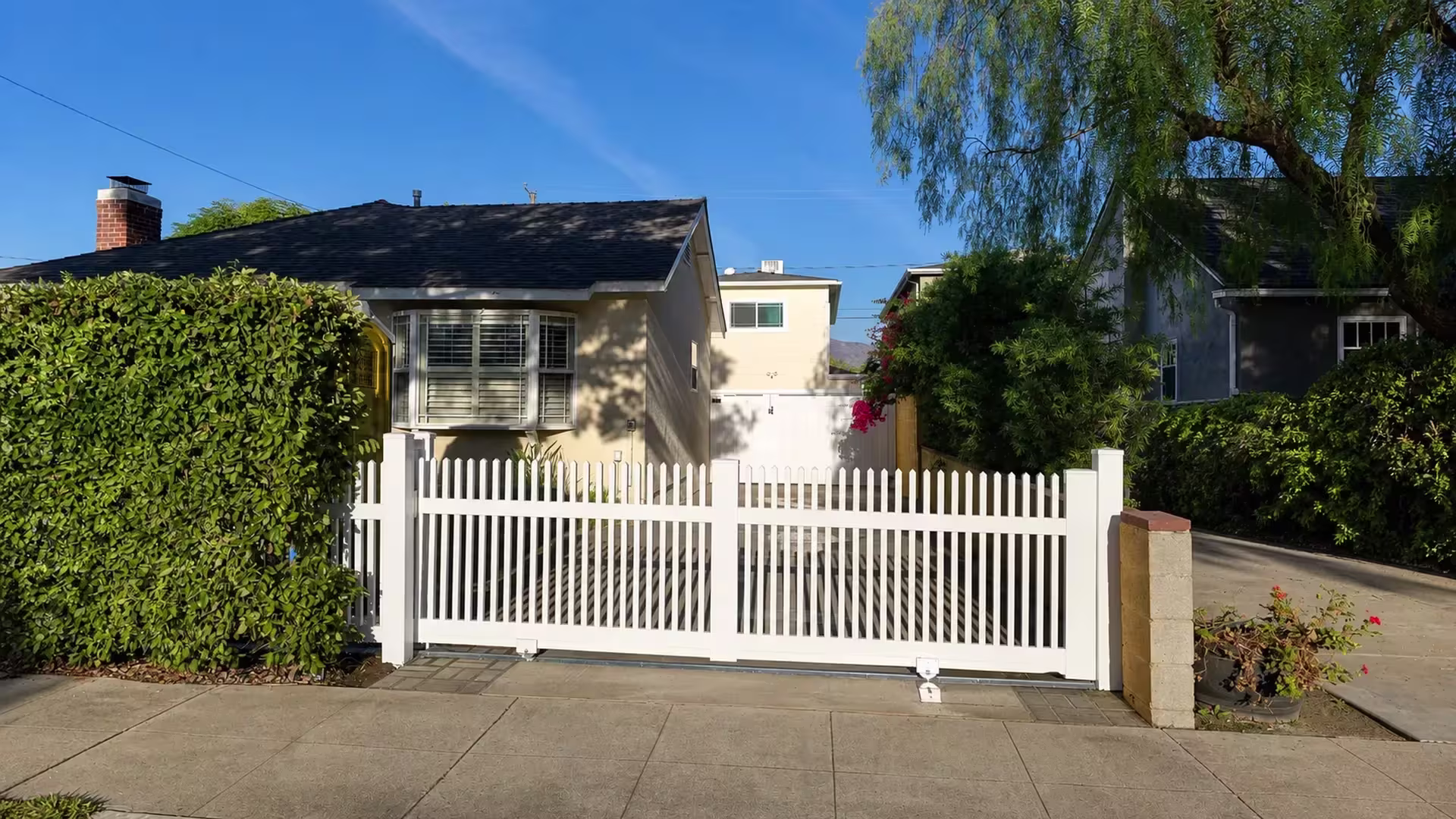 White vinyl picket-topped rolling gate sliding past a trimmed hedge toward a cream house and rear structure