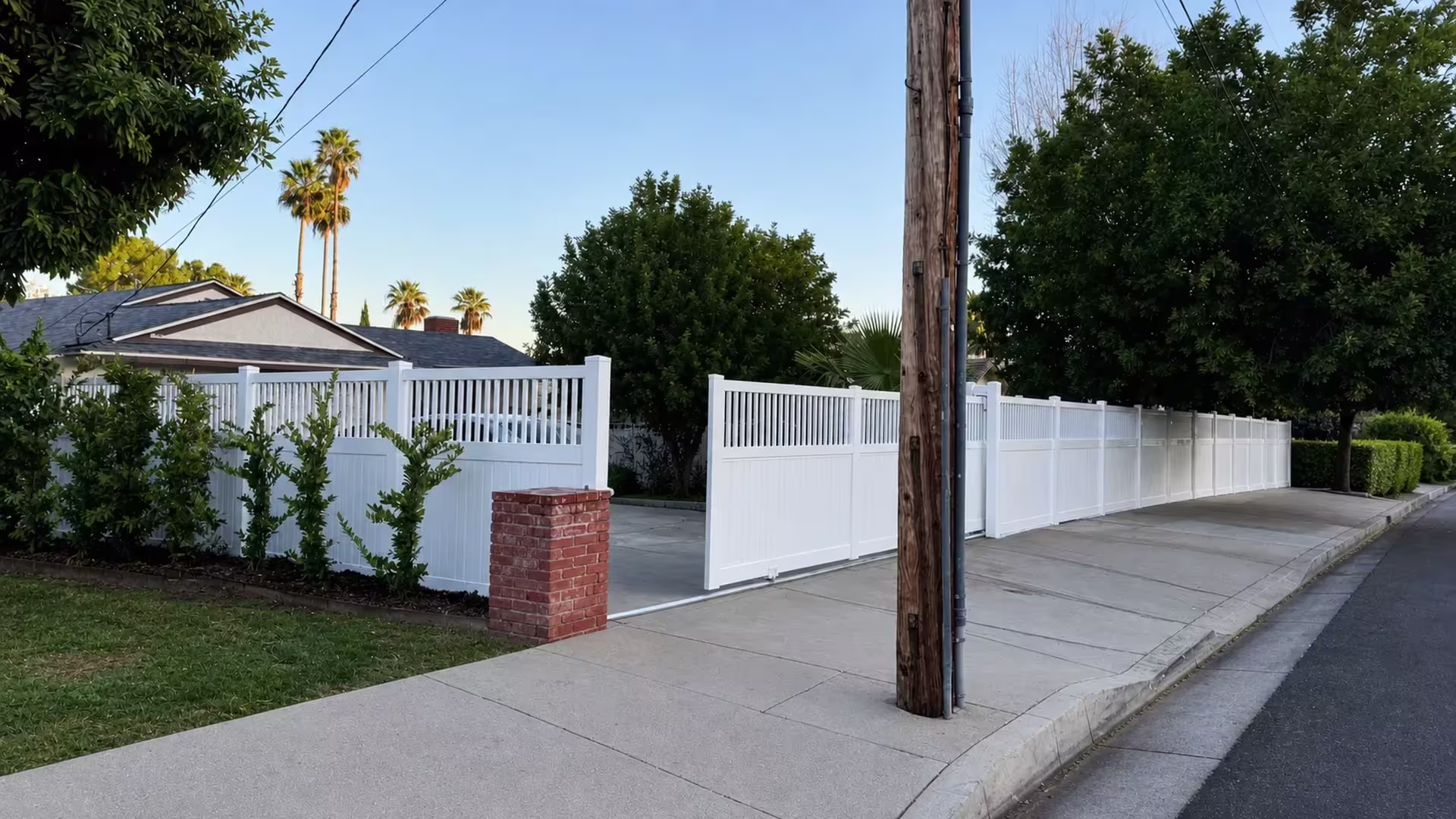 White vinyl picket-topped privacy rolling gate shown partly open beside a brick pillar under overhead utility wires