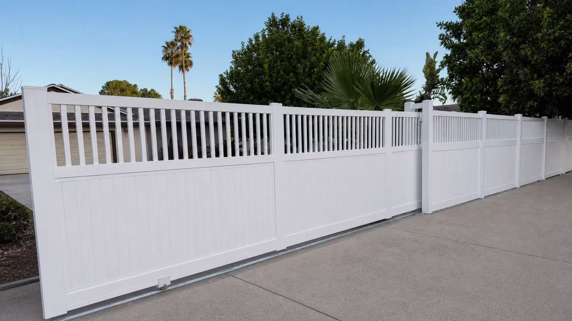 White vinyl privacy rolling gate with a picket top, its narrow end bay set beside mature fan palms