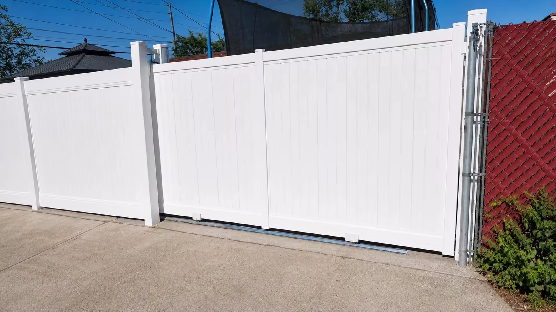 White vinyl privacy rolling gate sliding past a chain-link fence with red screening and climbing vines