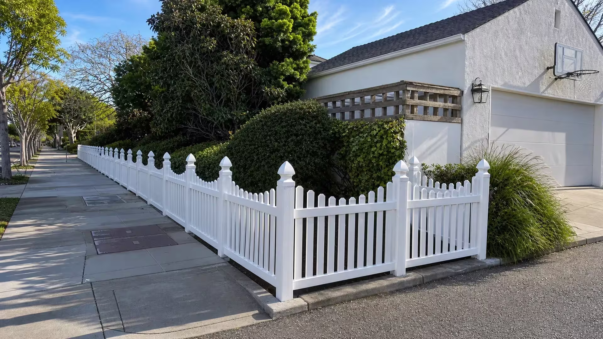 A weathered wood lattice screen stands near this White vinyl scalloped picket fence cornering the stucco house yard