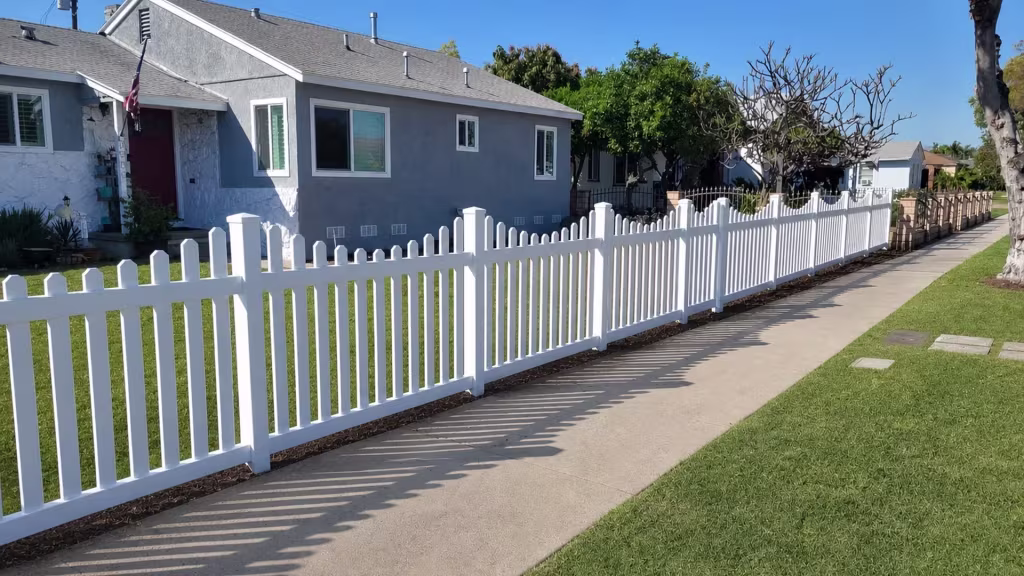 Picket tops dipping toward each panel center trace the rhythm of this White vinyl scalloped picket fence beside the sidewalk