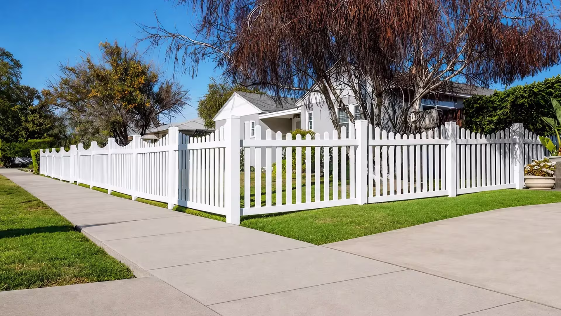 A square corner post anchors this White vinyl scalloped picket fence enclosing the front lawn near a planter