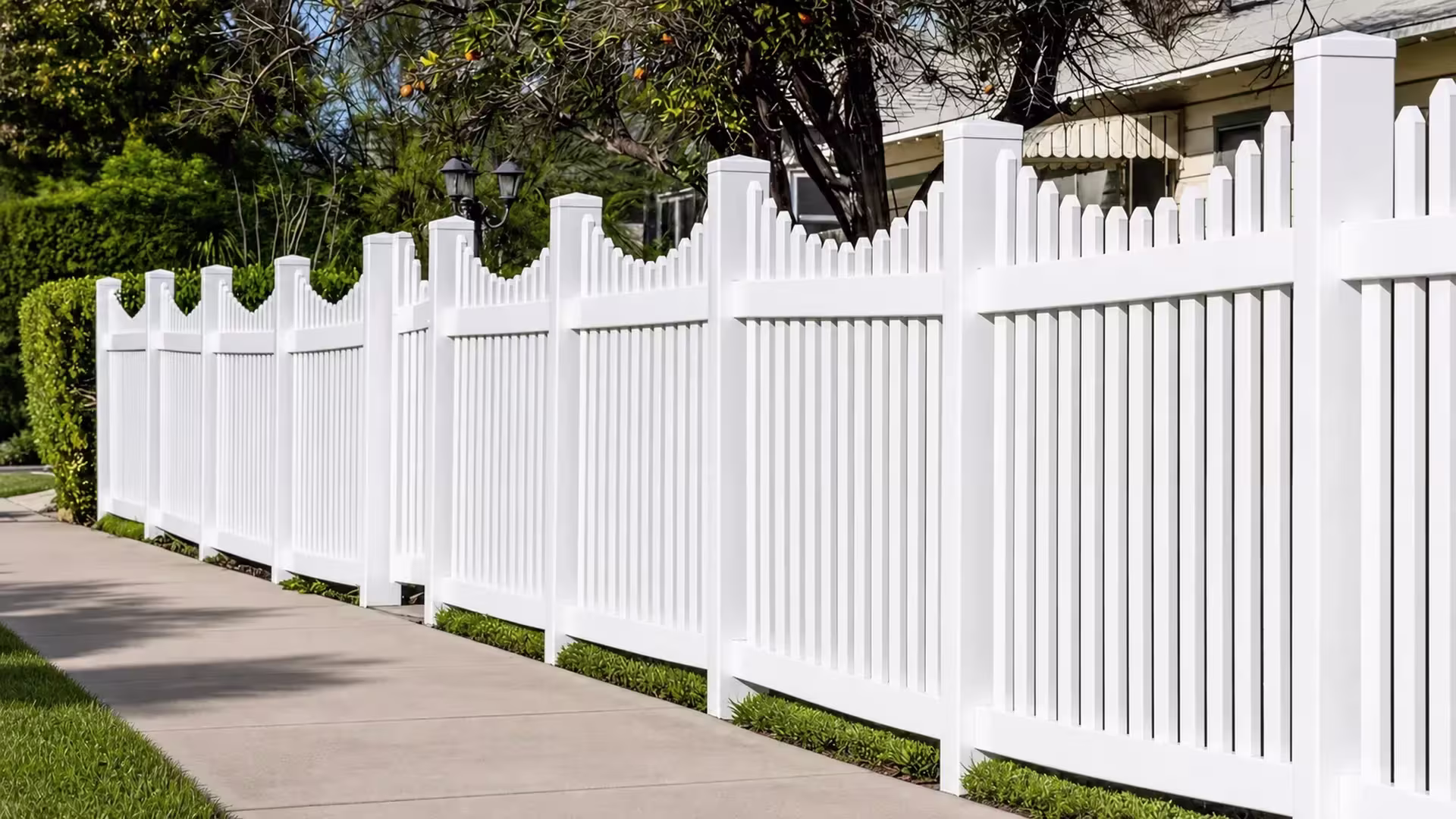 Twin garden lamps flank this White vinyl scalloped picket fence running past a cream stucco facade
