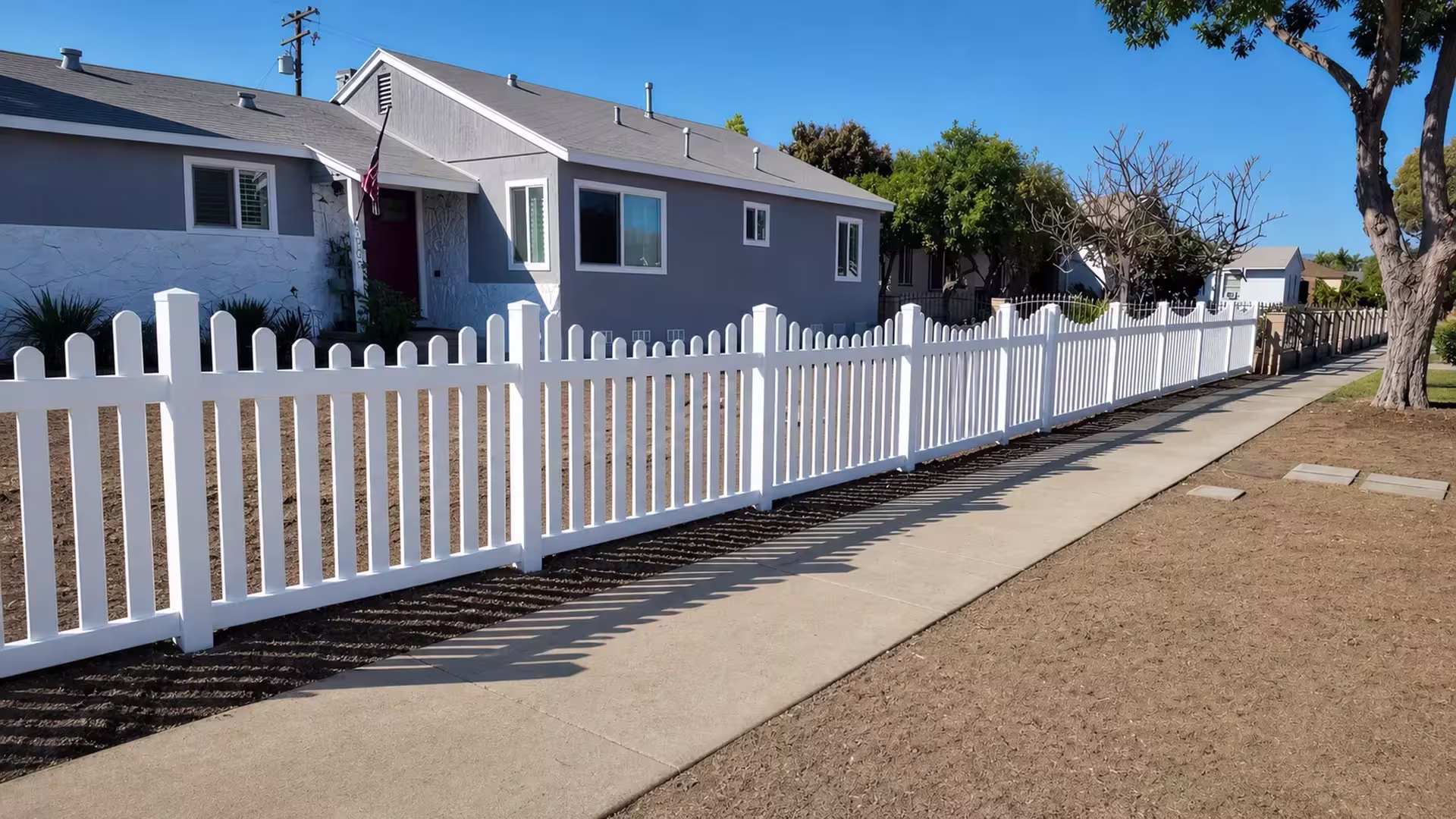 A dry graded yard stretches beside this White vinyl scalloped picket fence beneath overhead utility wires
