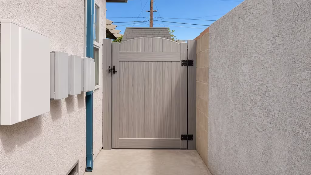 Mesquite vinyl side gate mounted beside a blue drainpipe and utility conduits on a stucco wall