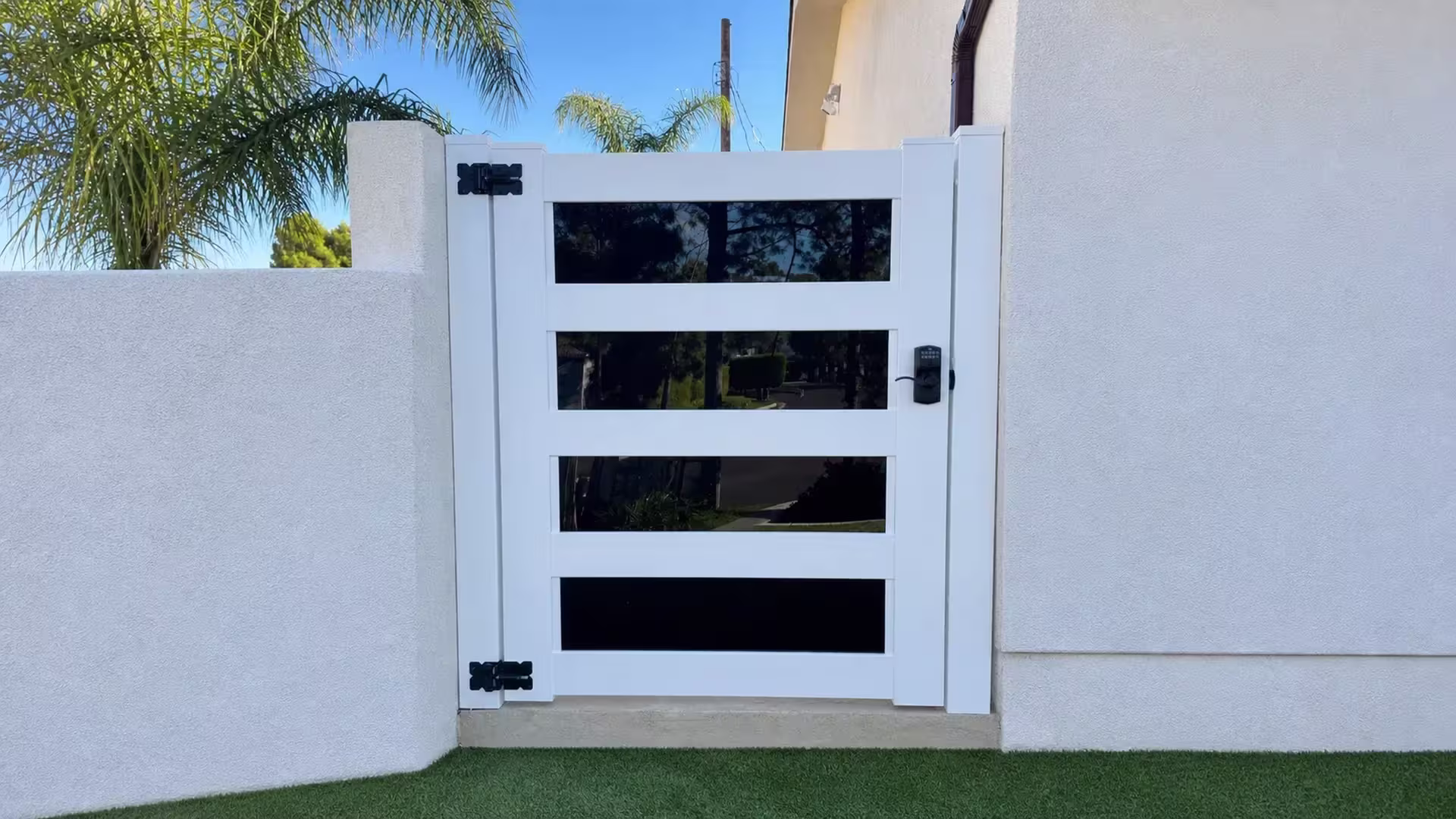 White vinyl side gate set between stucco walls beside artificial turf and tall palm trees