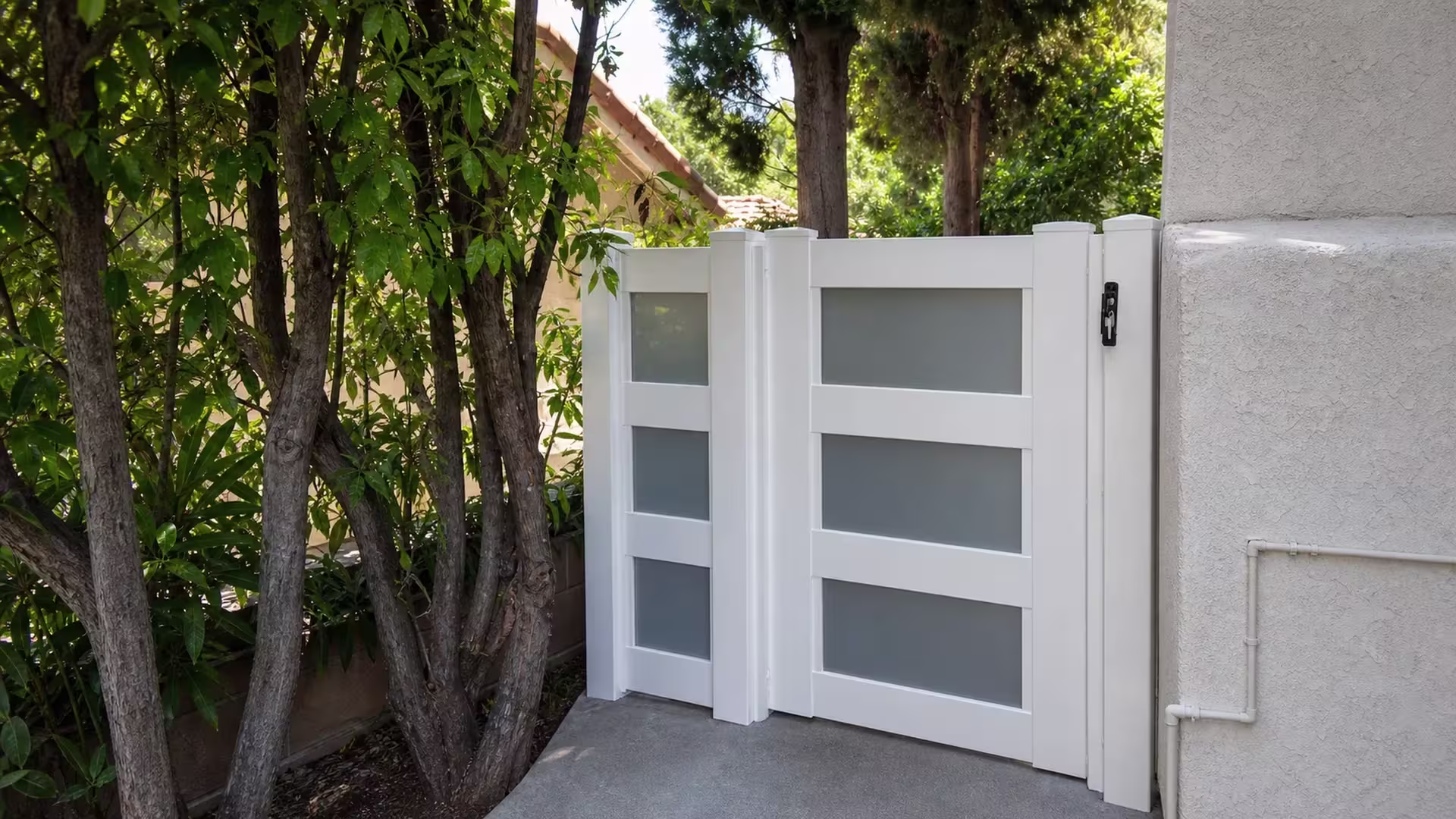 Mature trees flank this White vinyl side gate standing beside a light stucco building wall
