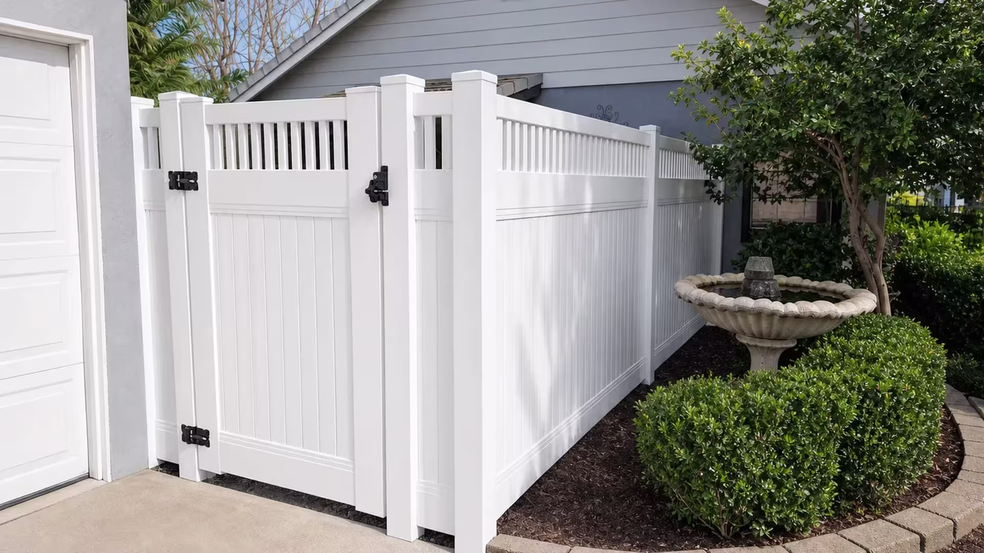 A picket top crowns this White vinyl side gate standing near a garage wall and a stone fountain