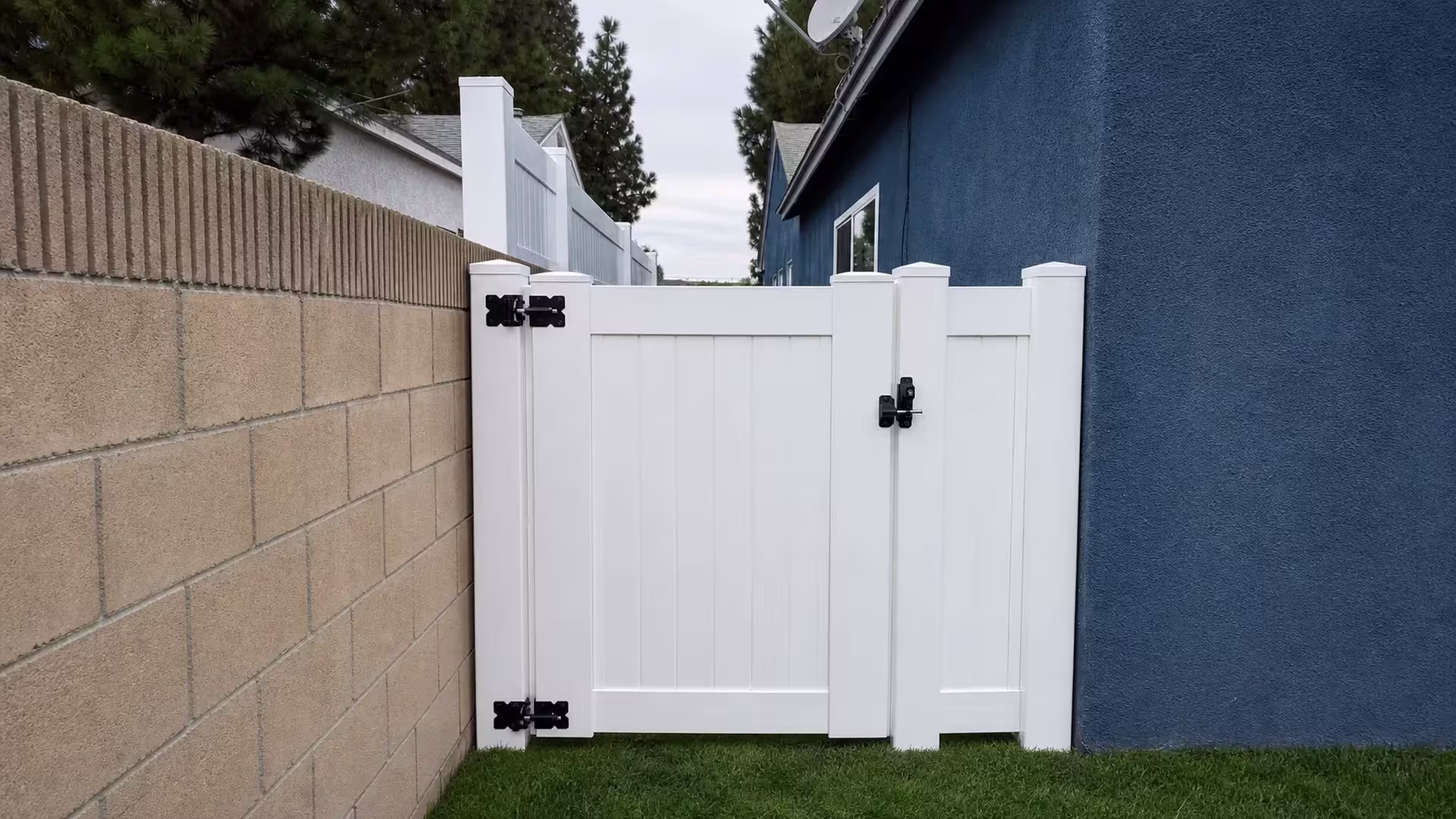 A narrow fixed panel joins this White vinyl side gate between a tan block wall and a dark blue stucco house