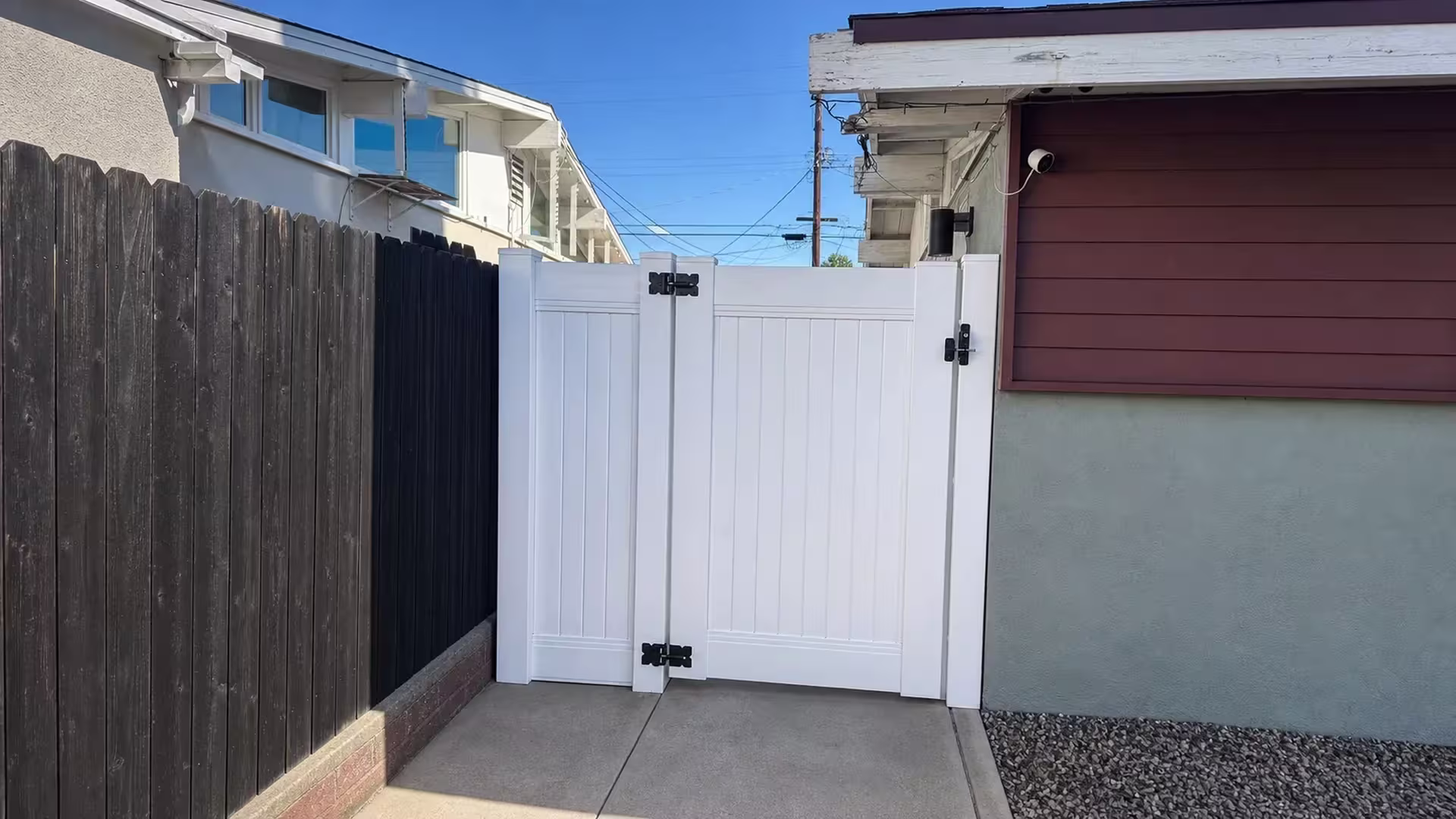 Two buildings frame this White vinyl side gate, paired with a narrower fixed panel of uneven width beside a black fence