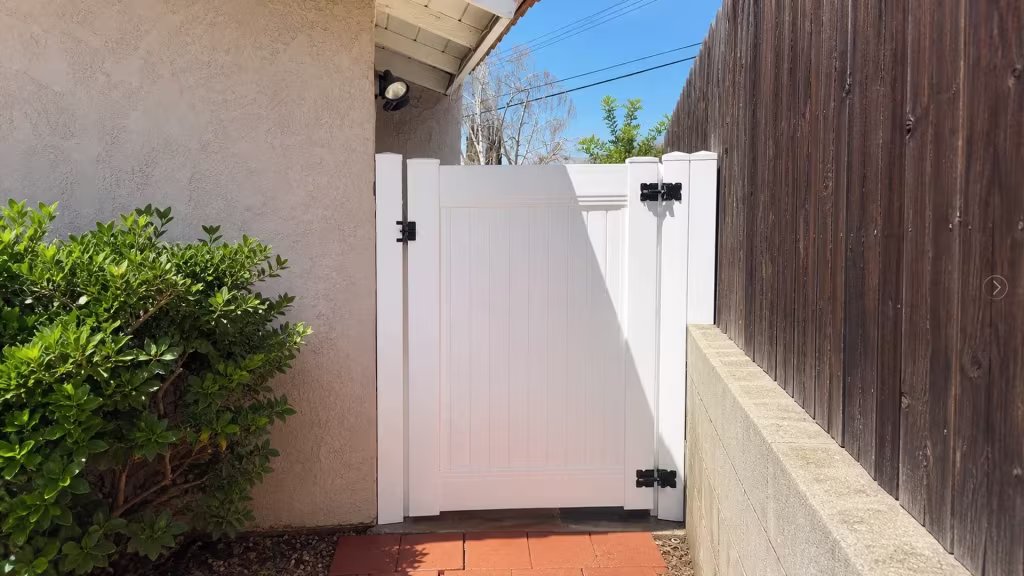 Red terracotta pavers lead to this White vinyl side gate, hinged at the right beside a weathered wood boundary fence