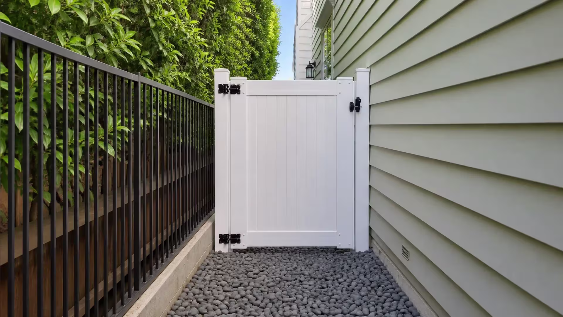 A black metal fence and pale green house siding flank this White vinyl side gate over river rock ground