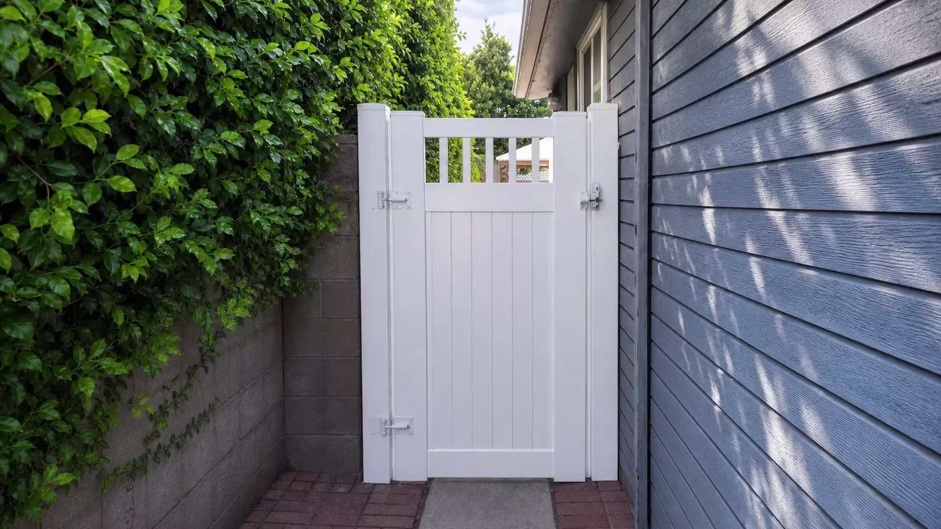 A masonry boundary wall and gray-blue wood siding border this White vinyl side gate along a brick walkway