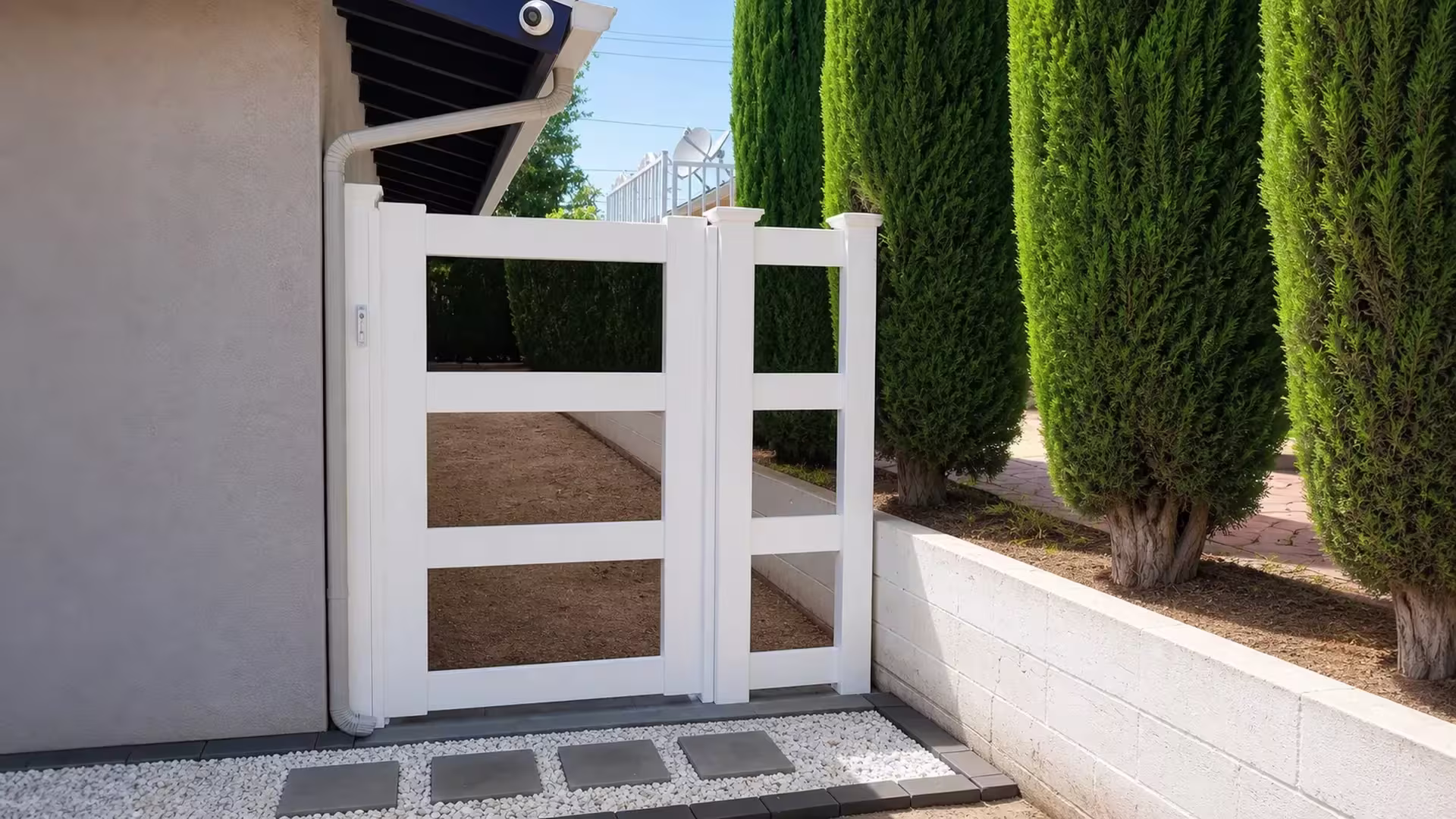 A stucco house and a low masonry wall lined with cypress flank this White vinyl side gate's asymmetrical two-leaf layout