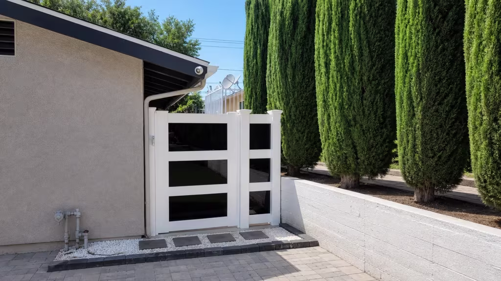 A center post divides this White vinyl side gate into a single passage beside a low masonry wall and cypress trees