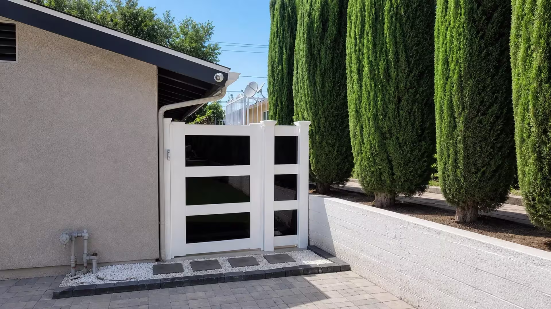 A center post divides this White vinyl side gate into a single passage beside a low masonry wall and cypress trees