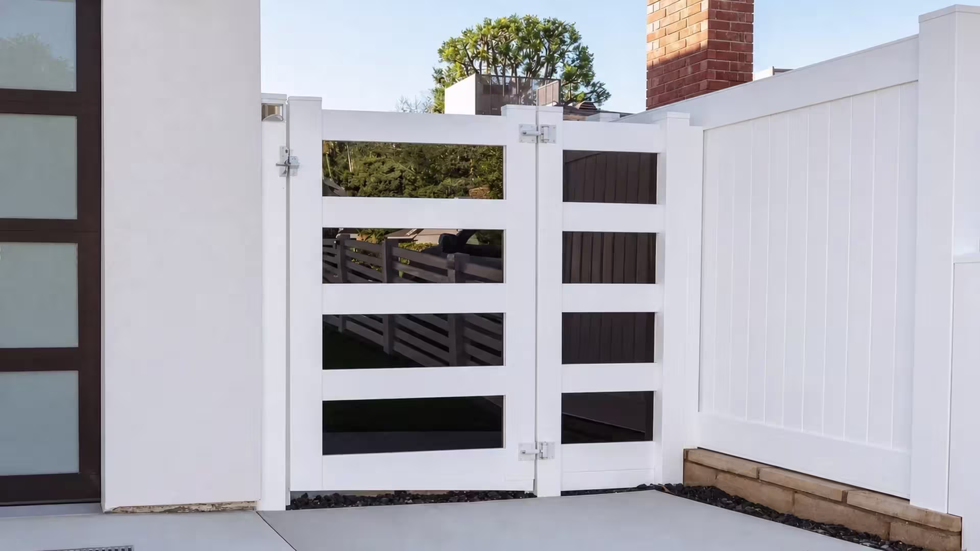 A brick chimney rises beside this White vinyl side gate hung between a house wall and a privacy fence