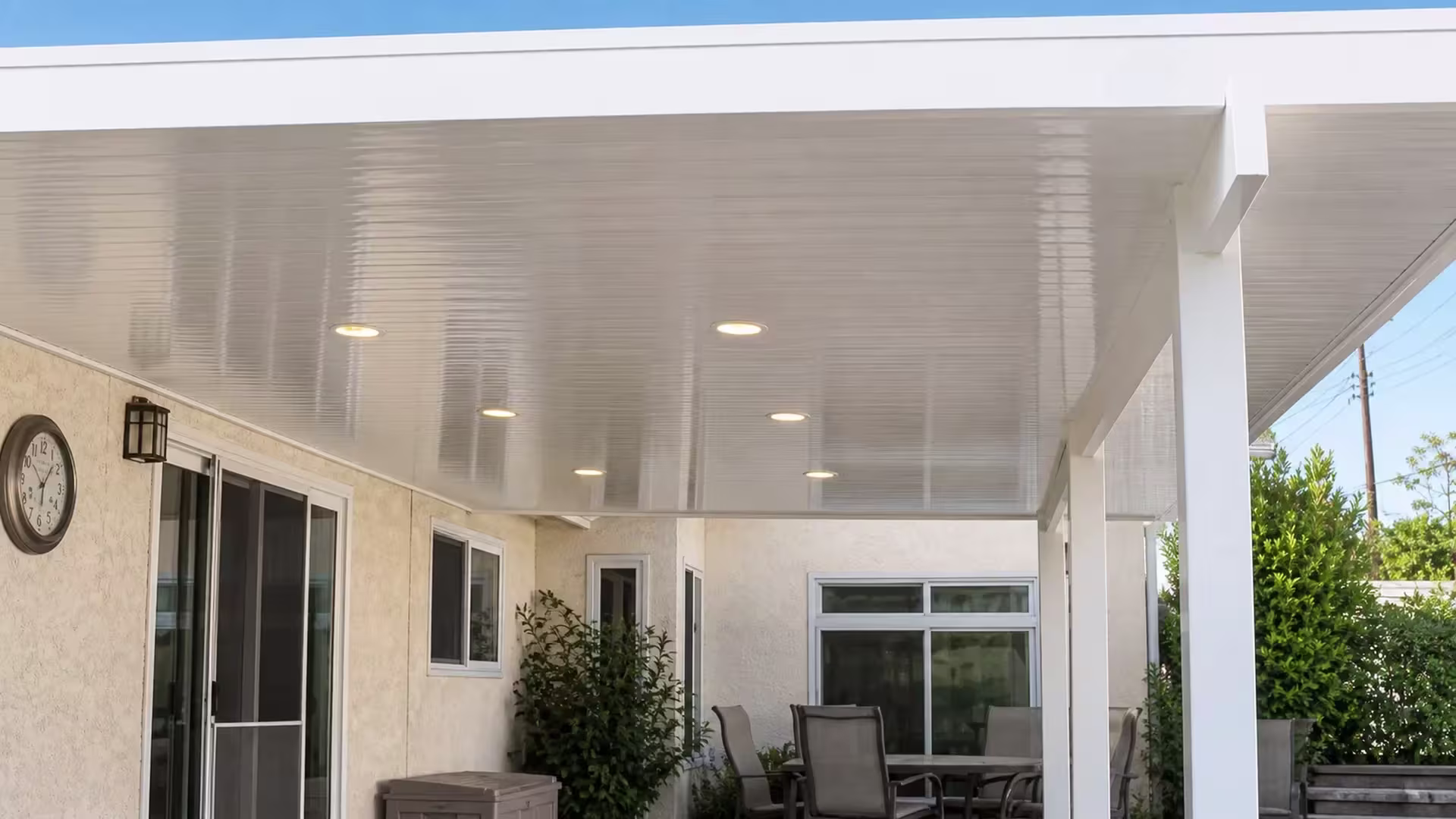 A wall clock hangs near the illuminated recessed lights beneath the White vinyl cover on beige stucco siding