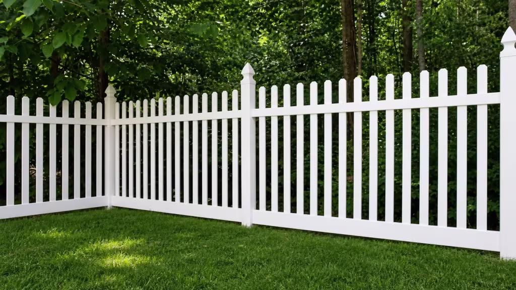 Dense shaded woodland trunks rise close behind the White vinyl picket fence at a shallow corner