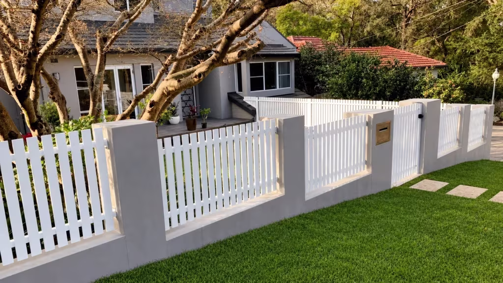 White vinyl picket fence sits above a low light gray masonry wall near a mailbox and lamp