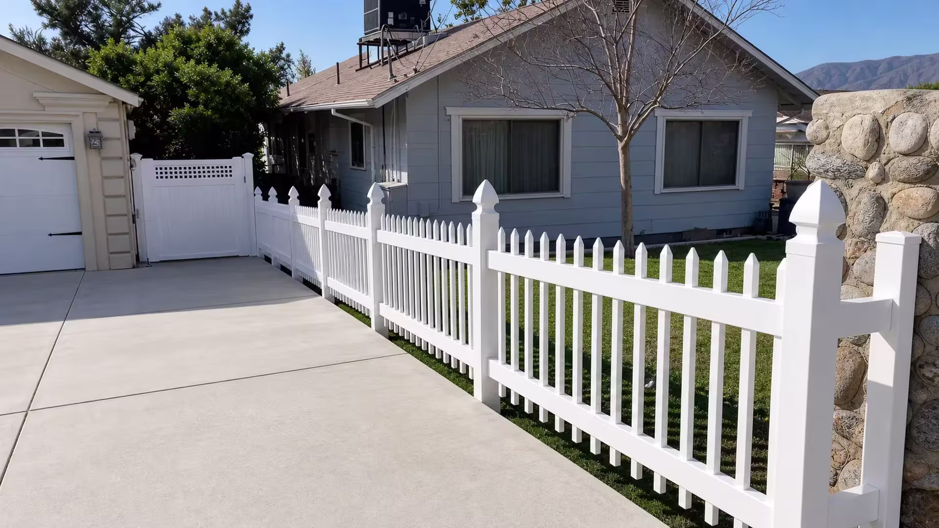 A diagonal run of White vinyl picket fence follows the concrete driveway past a sculpted rock wall