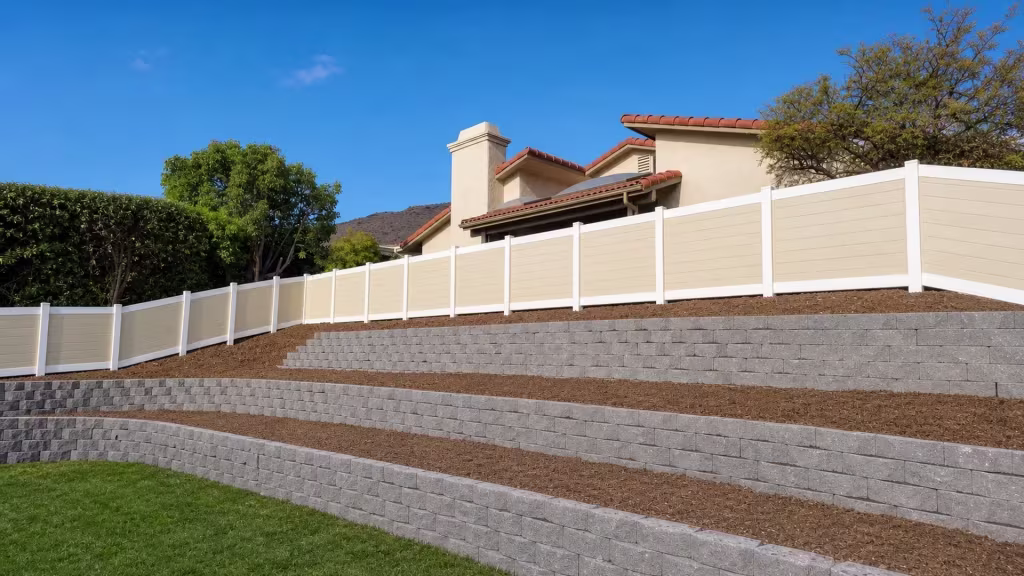 Beige vinyl two-tone fence climbing above terraced concrete-block retaining walls on a hillside