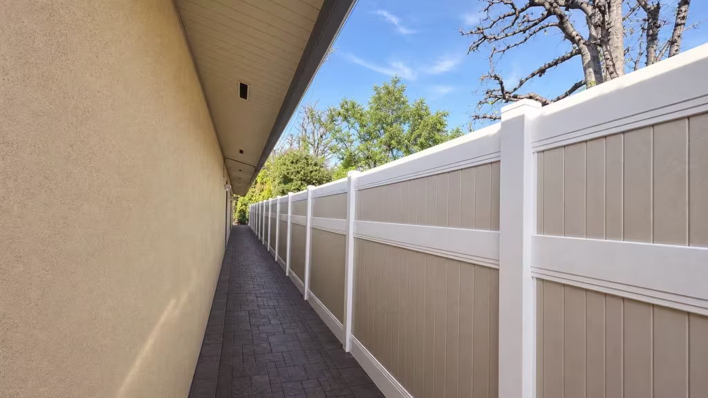 Mature trees rise beyond the stucco wall where a White vinyl two-tone fence lines the paver walk