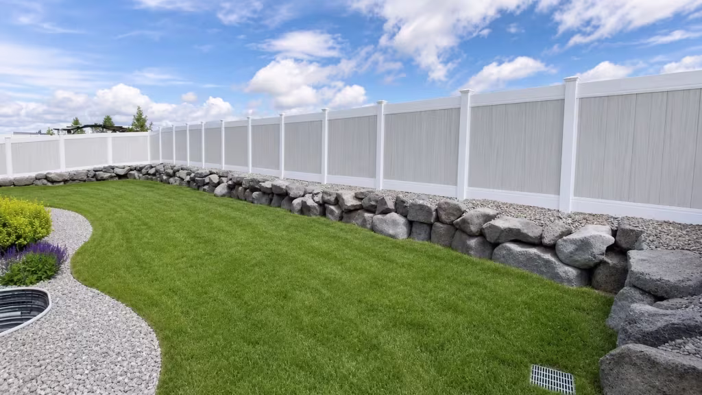 Vinyl two-tone privacy fence turning a curved lawn boundary past a low boulder edge and gravel beds