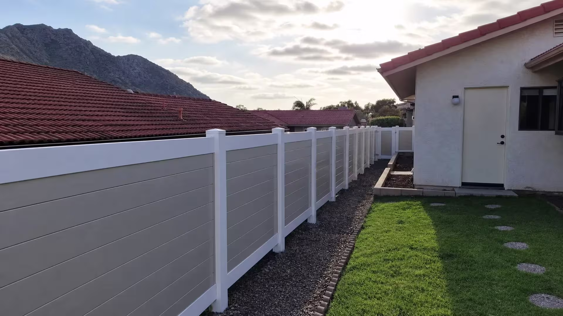 Red tile roofs and distant mountains rise behind a White vinyl two-tone privacy fence