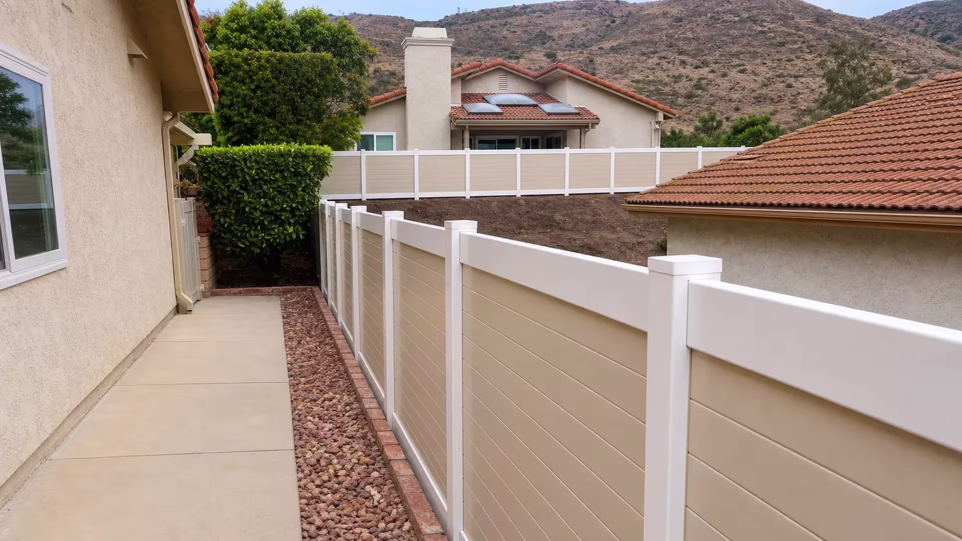 A narrow concrete walkway runs beside a Tan vinyl two-tone horizontal privacy fence and a rock bed