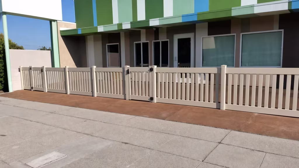 A Clay vinyl closed-top picket fence fronts a commercial storefront, six posts with beveled caps under a striped awning