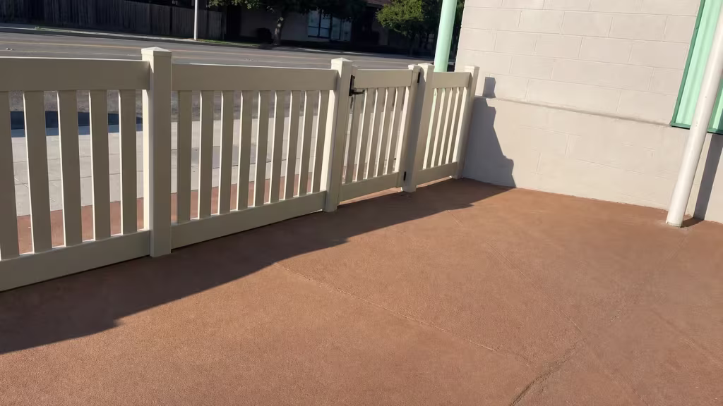 A beige block wall runs beside a Tan vinyl fence of closed-top pickets angling toward a masonry building
